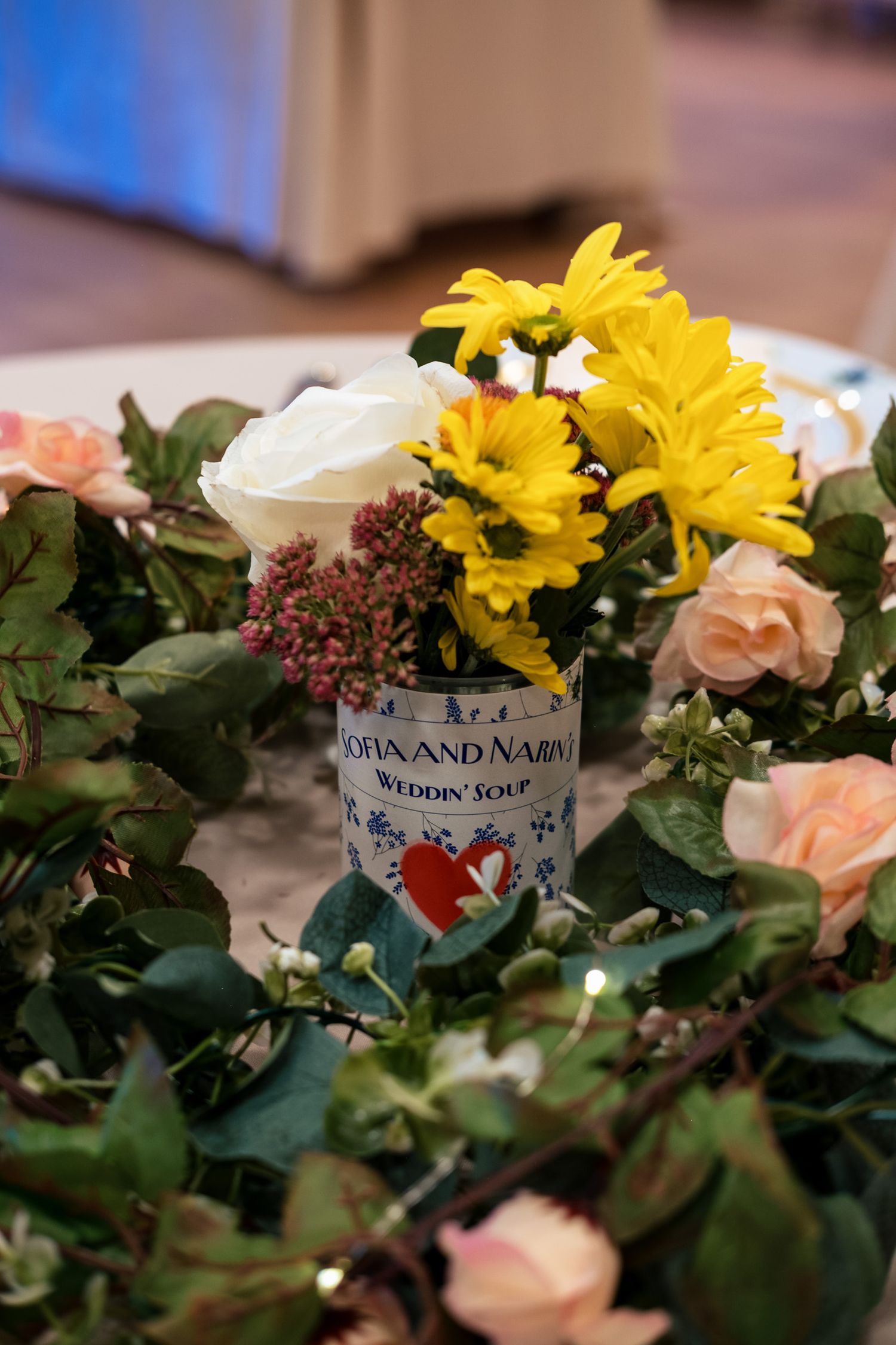 Sofia and Nabin wedding centerpiece featuring yellow daisies and white roses in a custom soup can surrounded by greenery.