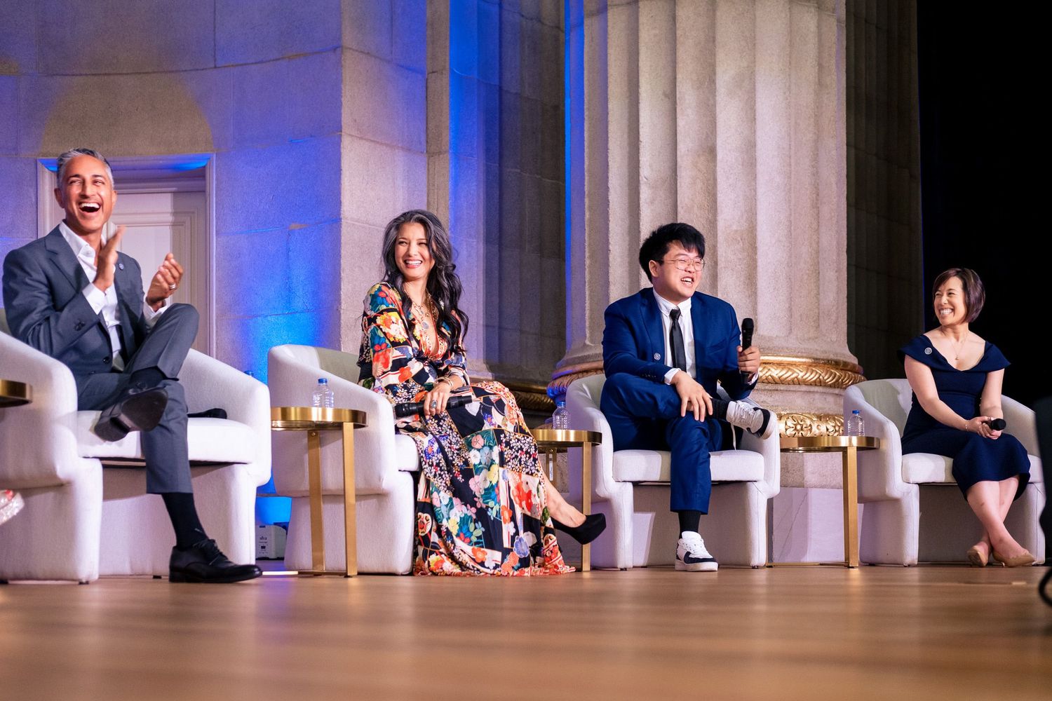 Four panelists seated on stage at a formal event, smiling and applauding in an elegant columned venue.
