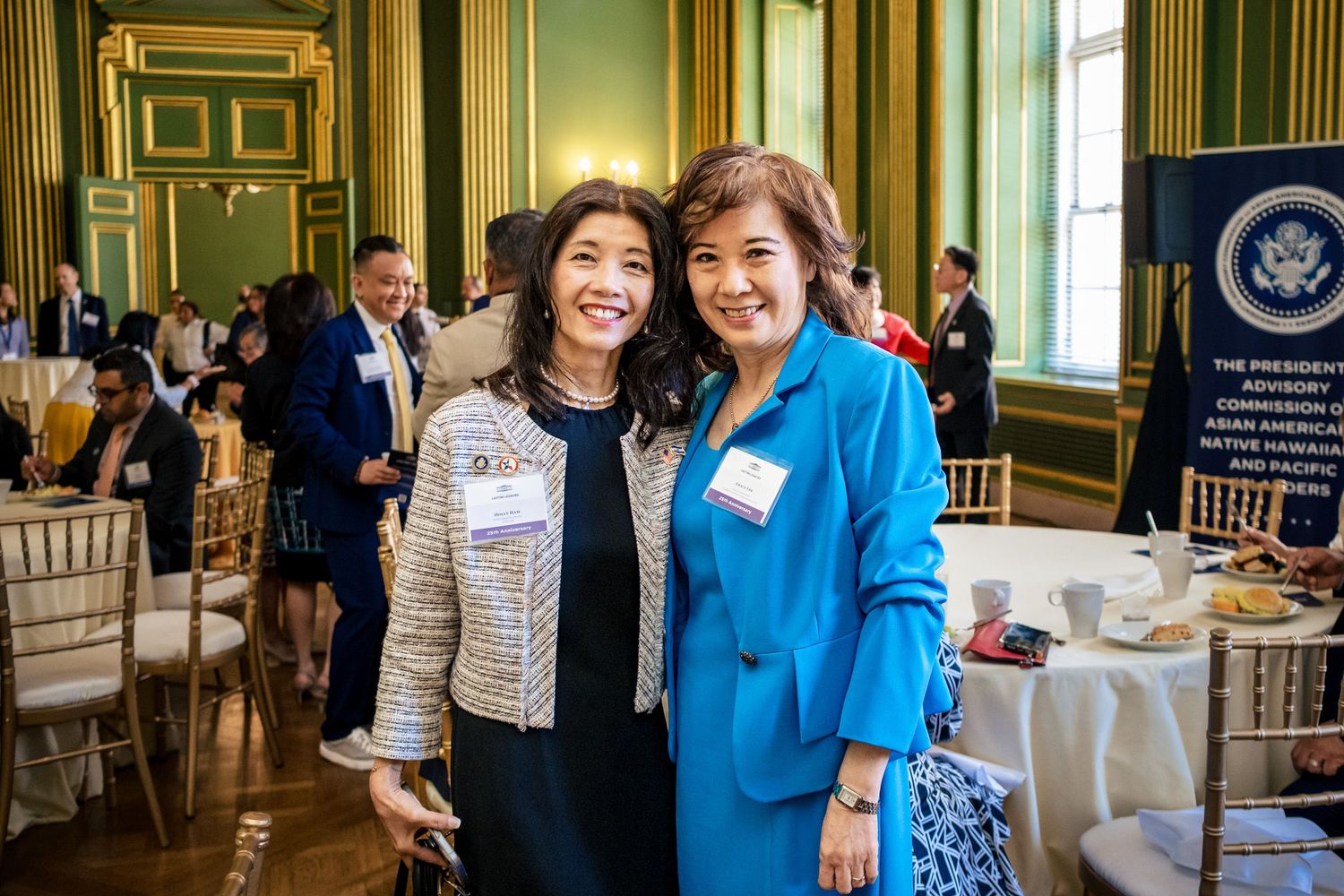 Two women wearing name badges smile together at a formal event in an elegant green ballroom.