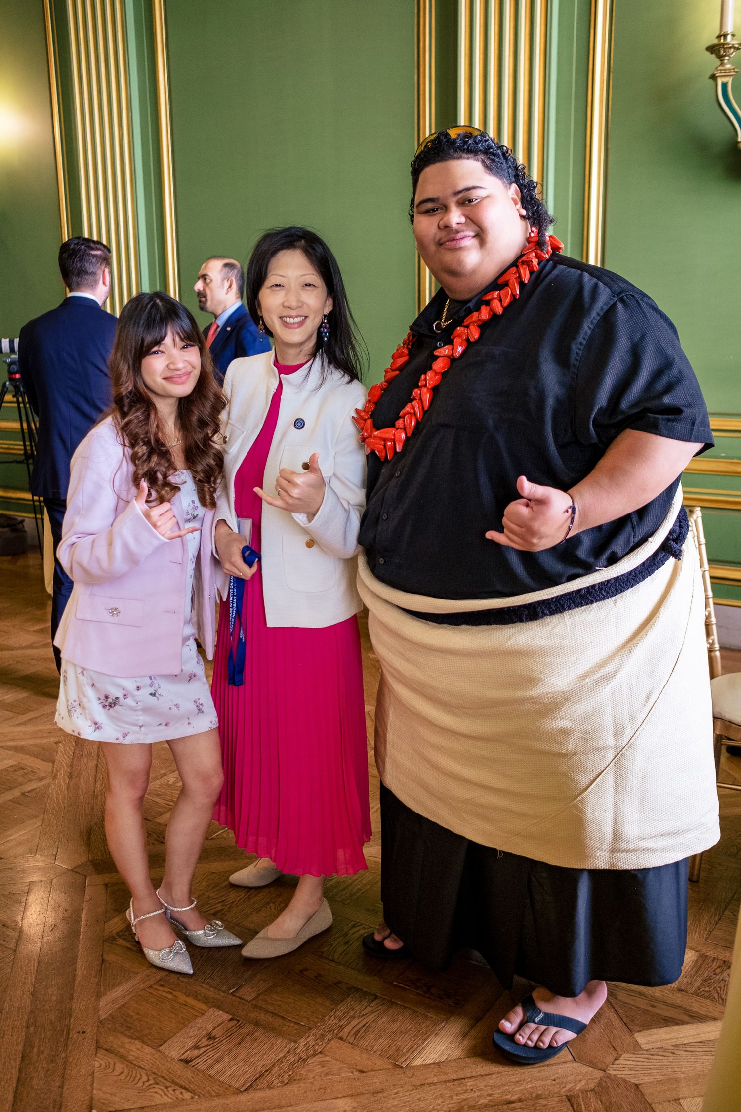 Three people posing together in an elegant green room, one wearing a traditional lei and cultural attire.