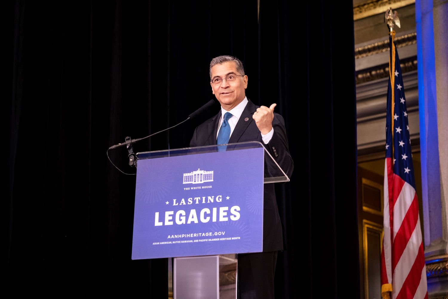 Man speaking at podium with Lasting Legacies sign, American flag backdrop, formal event setting.