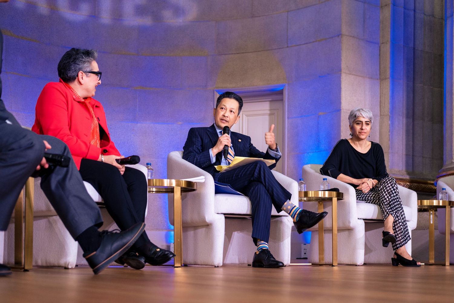 Three panelists seated on white chairs at a formal conference, with a man in a blue suit speaking animatedly on stage.
