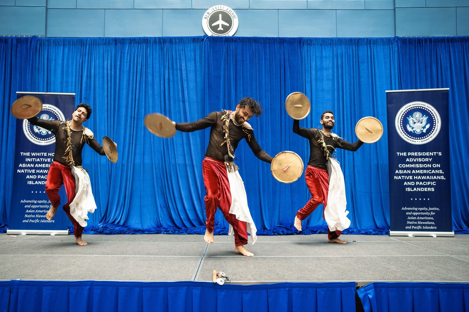 Three male dancers in traditional red and white costumes perform with cymbals on a blue-draped stage.