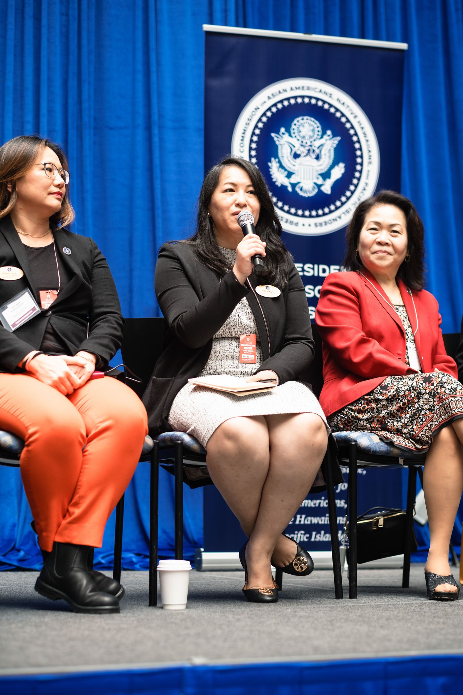 Three women seated on stage at a Presidential advisory council panel event with US government banner backdrop.