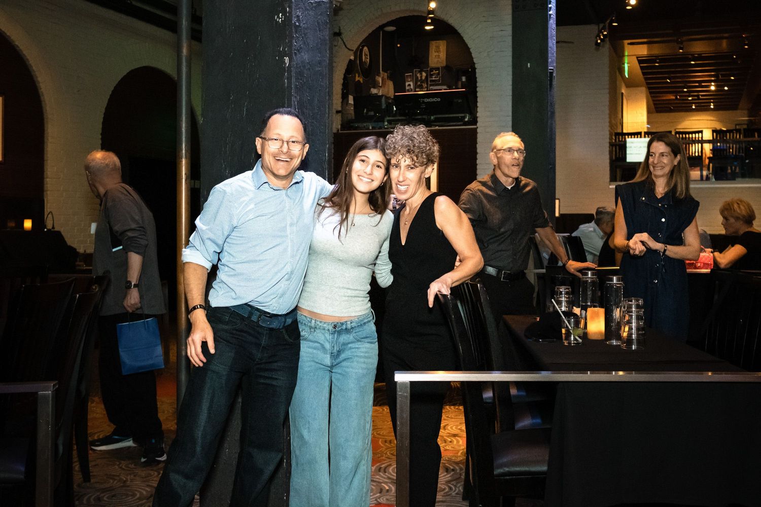 Three people smiling and posing together inside a dimly lit restaurant or bar setting.