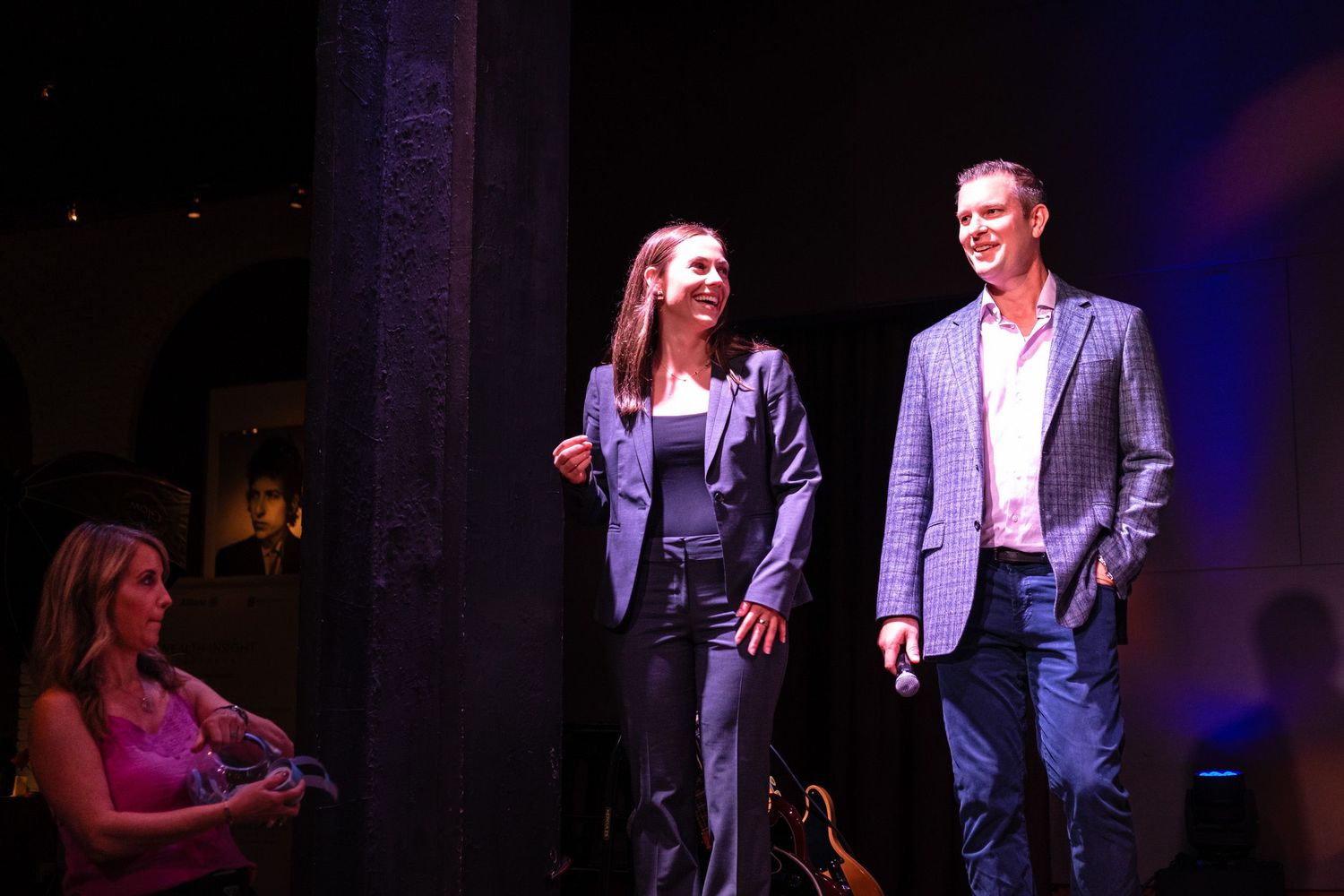 Man and woman presenting on stage at a formal evening event with audience in background.