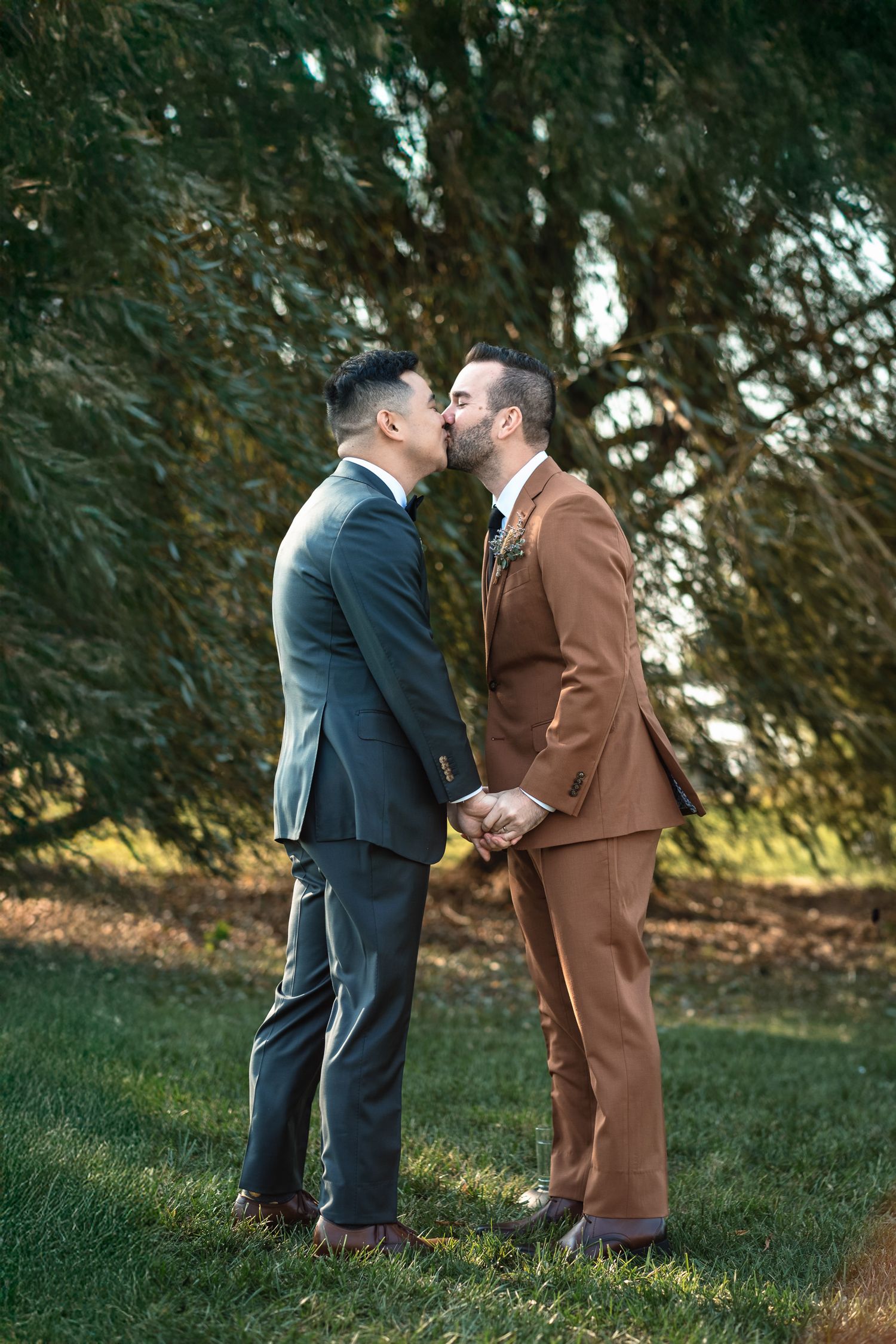 Two grooms share a first kiss as married couple during their outdoor wedding ceremony on green grass.