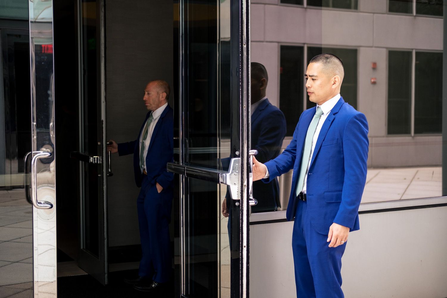 Groom in royal blue suit opens glass door of modern building on wedding day.