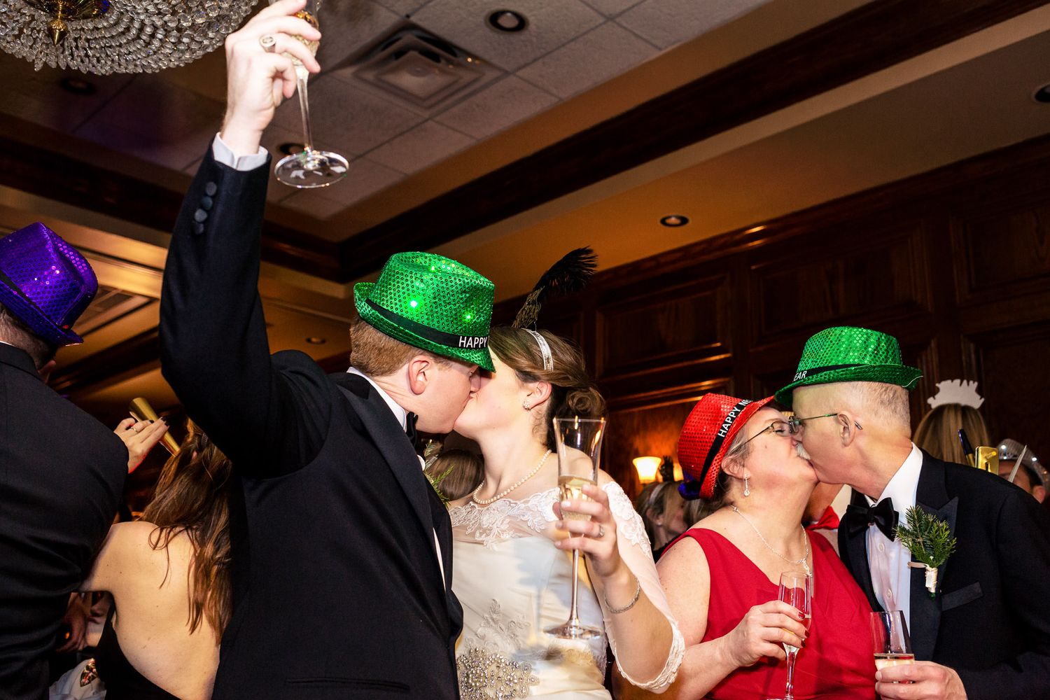 Two couples in green hats share New Year's Eve kisses while holding champagne glasses at a festive wedding reception.