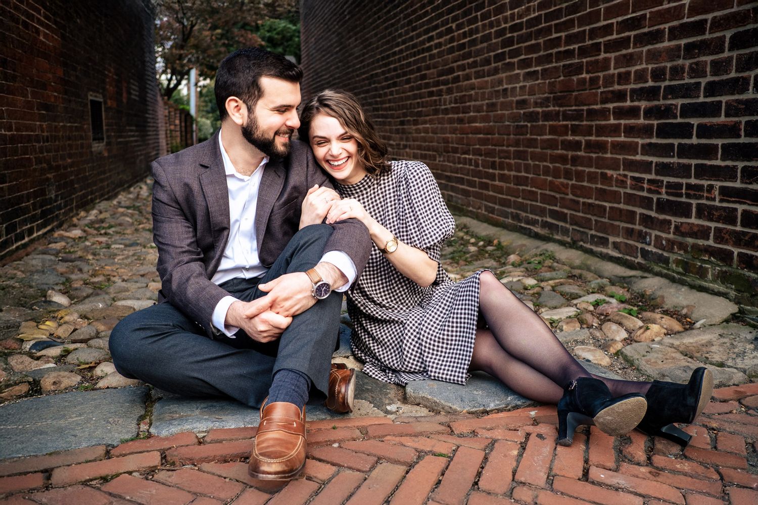 Happy couple sitting on brick cobblestone path, laughing and embracing in an urban alley.