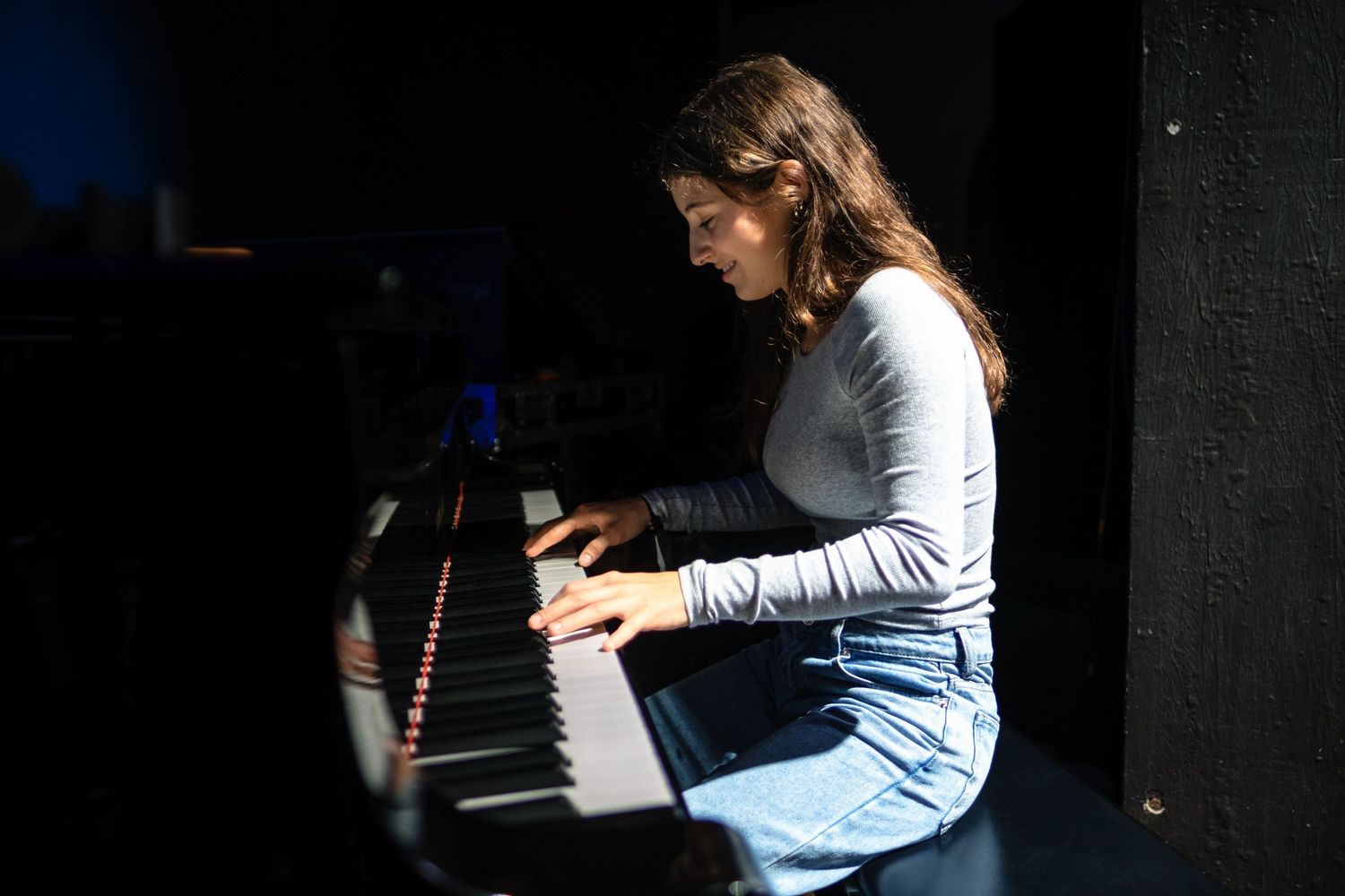 Young woman with brown hair playing a black piano in a dimly lit room, wearing a gray long-sleeve shirt.