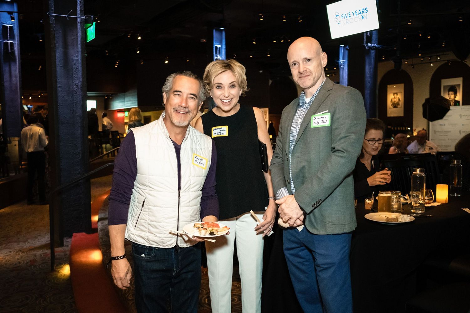 Three professionals smiling together at a corporate networking event in a dimly lit venue.