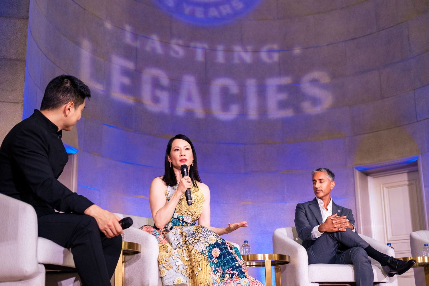 Three panelists discuss on stage at a Lasting Legacies event, with a woman in floral dress speaking into a microphone.