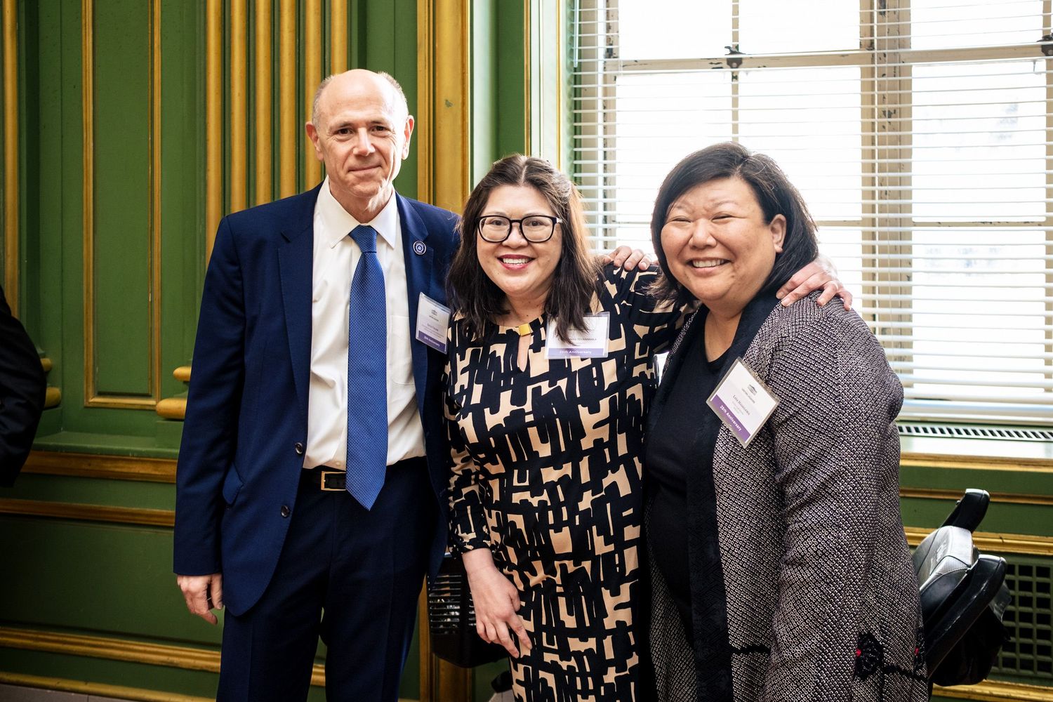 Three professionals smiling together at a formal event in an elegant green-walled room, wearing business attire and name badges.