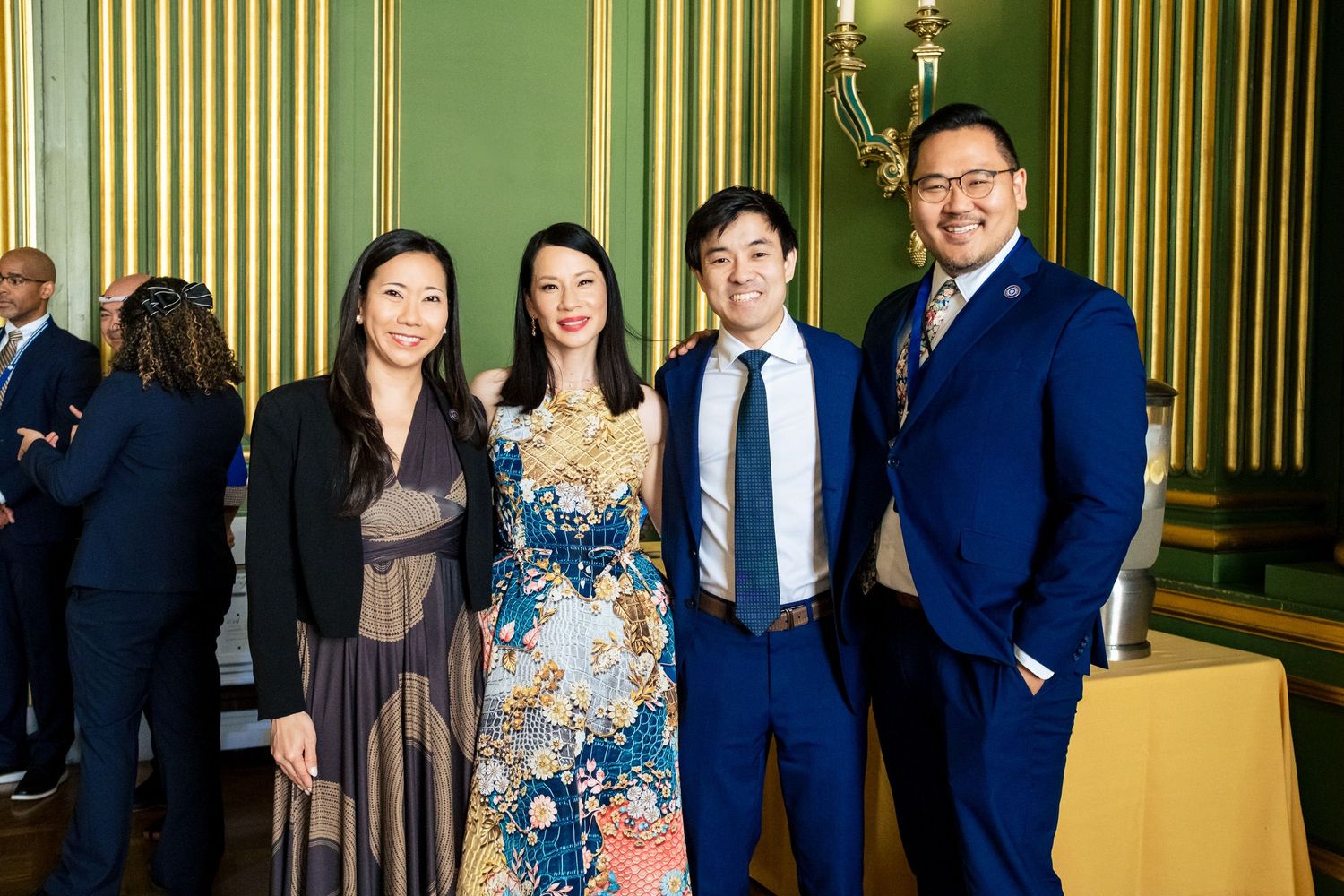 Four elegantly dressed people posing together in a green ornate room at a formal event.