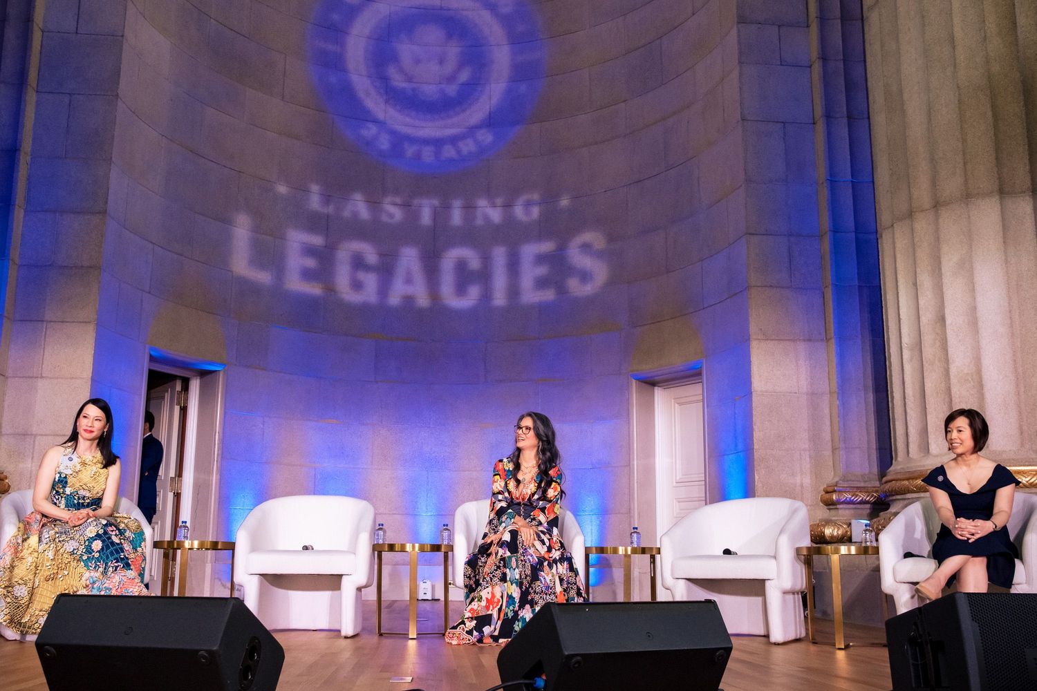 Three women on stage at Lasting Legacies event, seated in white chairs under blue lighting at elegant venue.