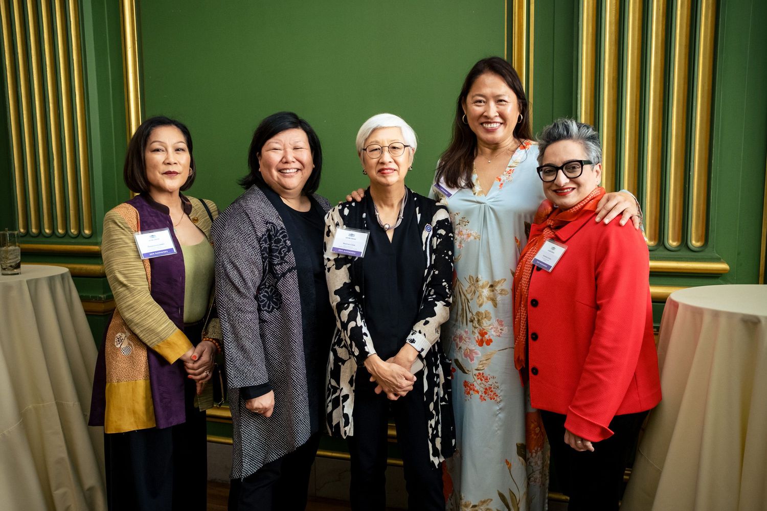 Five women smiling and posing together at a formal event in an elegant green-walled venue.