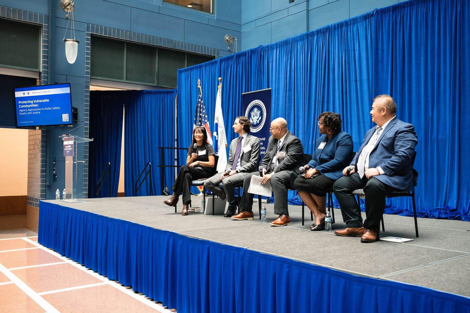 Five panelists seated on a blue-draped stage at a formal government event with US and agency flags.