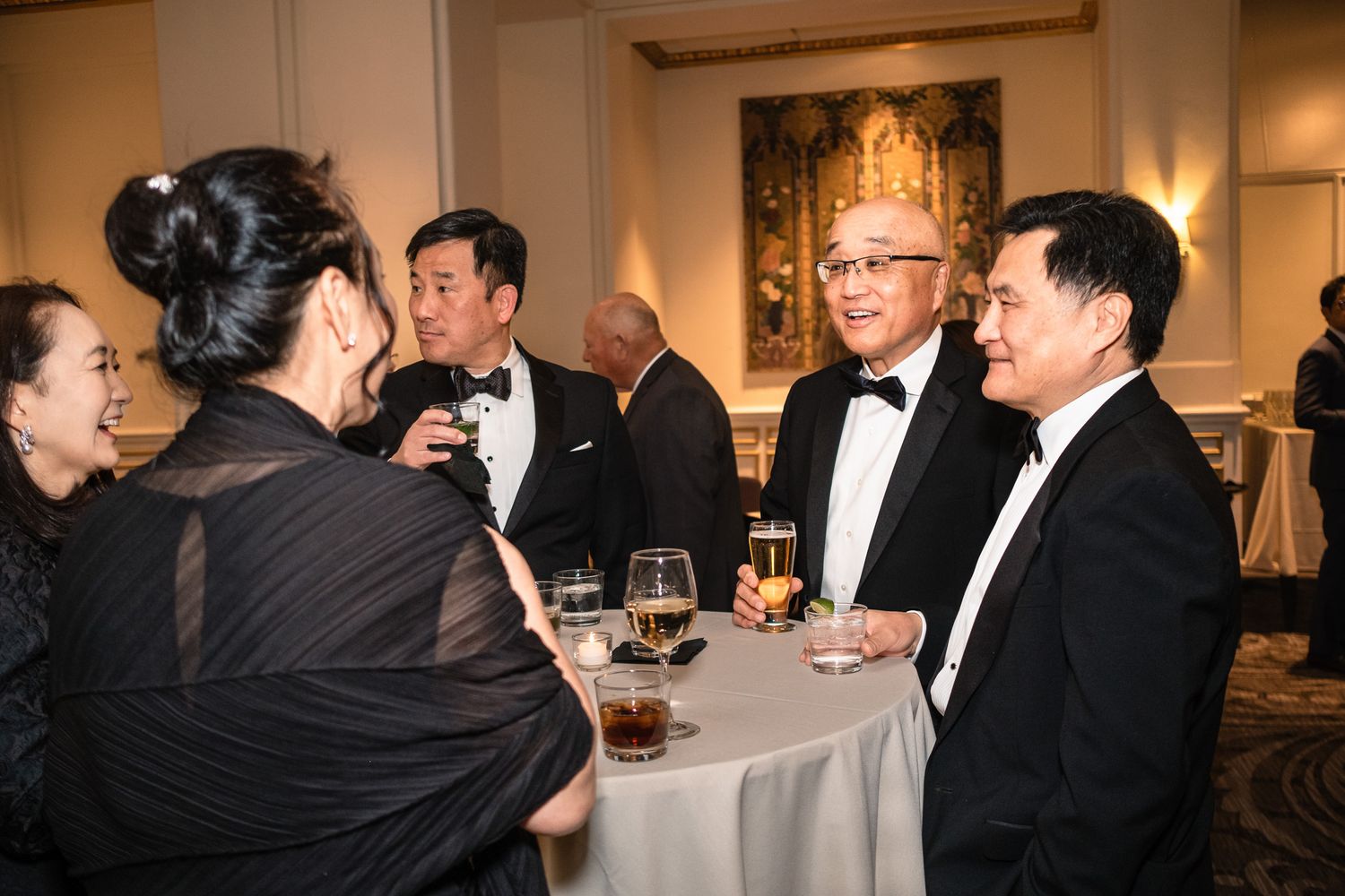 Elegantly dressed guests in formal attire socializing at a black tie gala event in a grand ballroom.