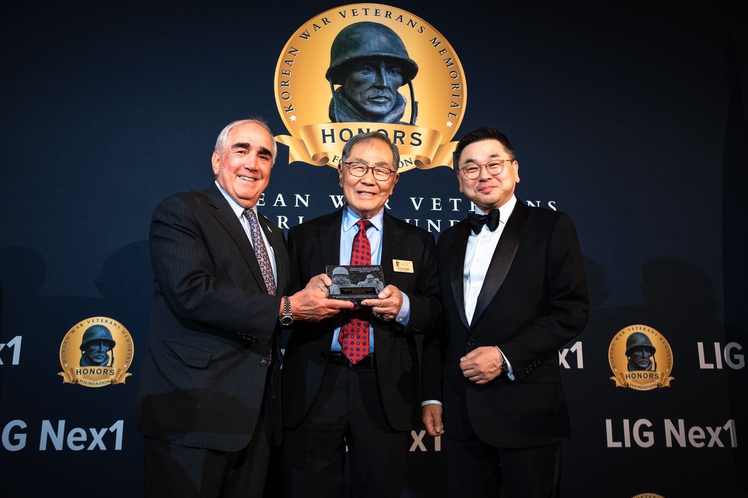 Three men pose together at Korean War Veterans Memorial Foundation honors ceremony, middle man holding award plaque.