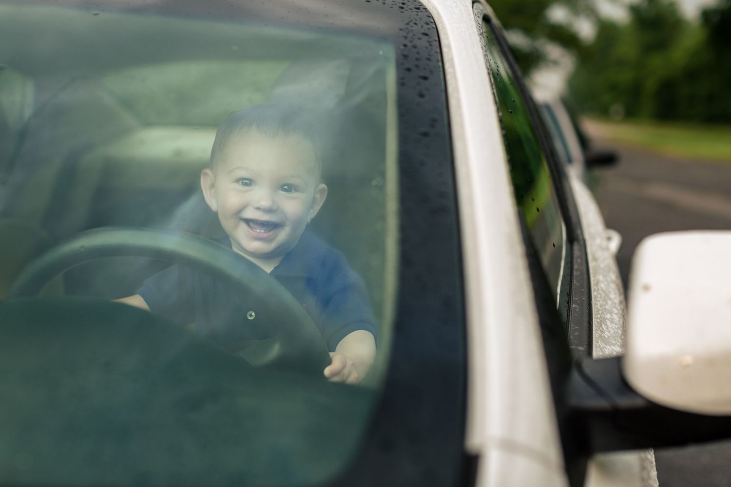Happy smiling baby sitting behind car steering wheel looking through windshield.