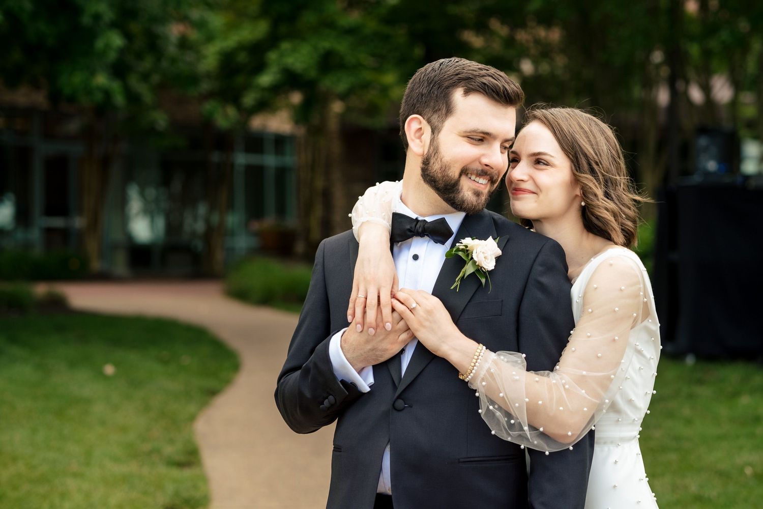 Couple embracing outdoors, groom in black tuxedo and bride in lace wedding gown.