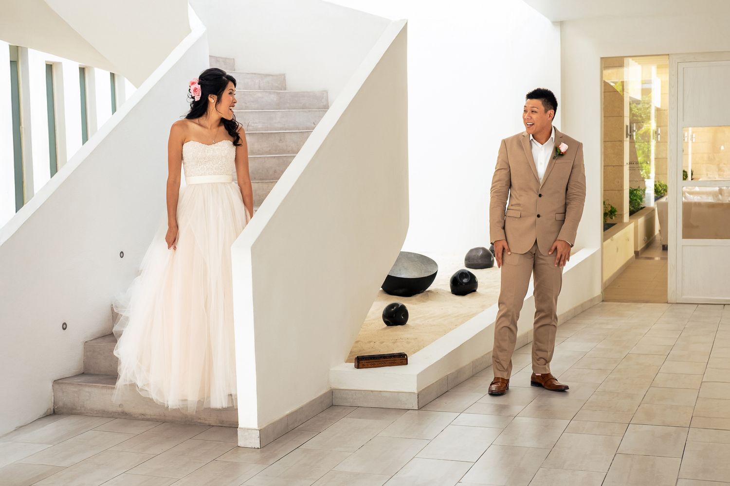 Bride in white gown and groom in tan suit share a first look moment in a modern white interior staircase.