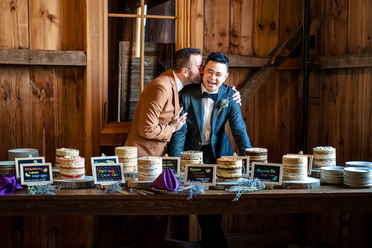 Two grooms share a joyful embrace beside a rustic pie display table at their wedding.