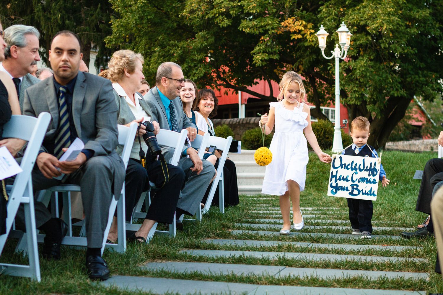 Young flower girl walks down outdoor aisle holding sign for wedding ceremony guests.