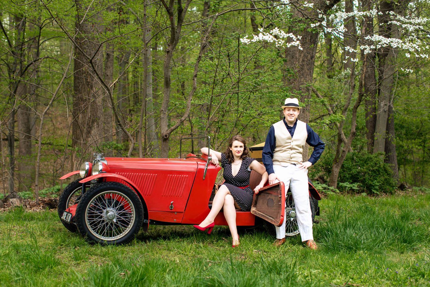Elegant couple posing beside a classic red vintage car on a green meadow in spring woods.