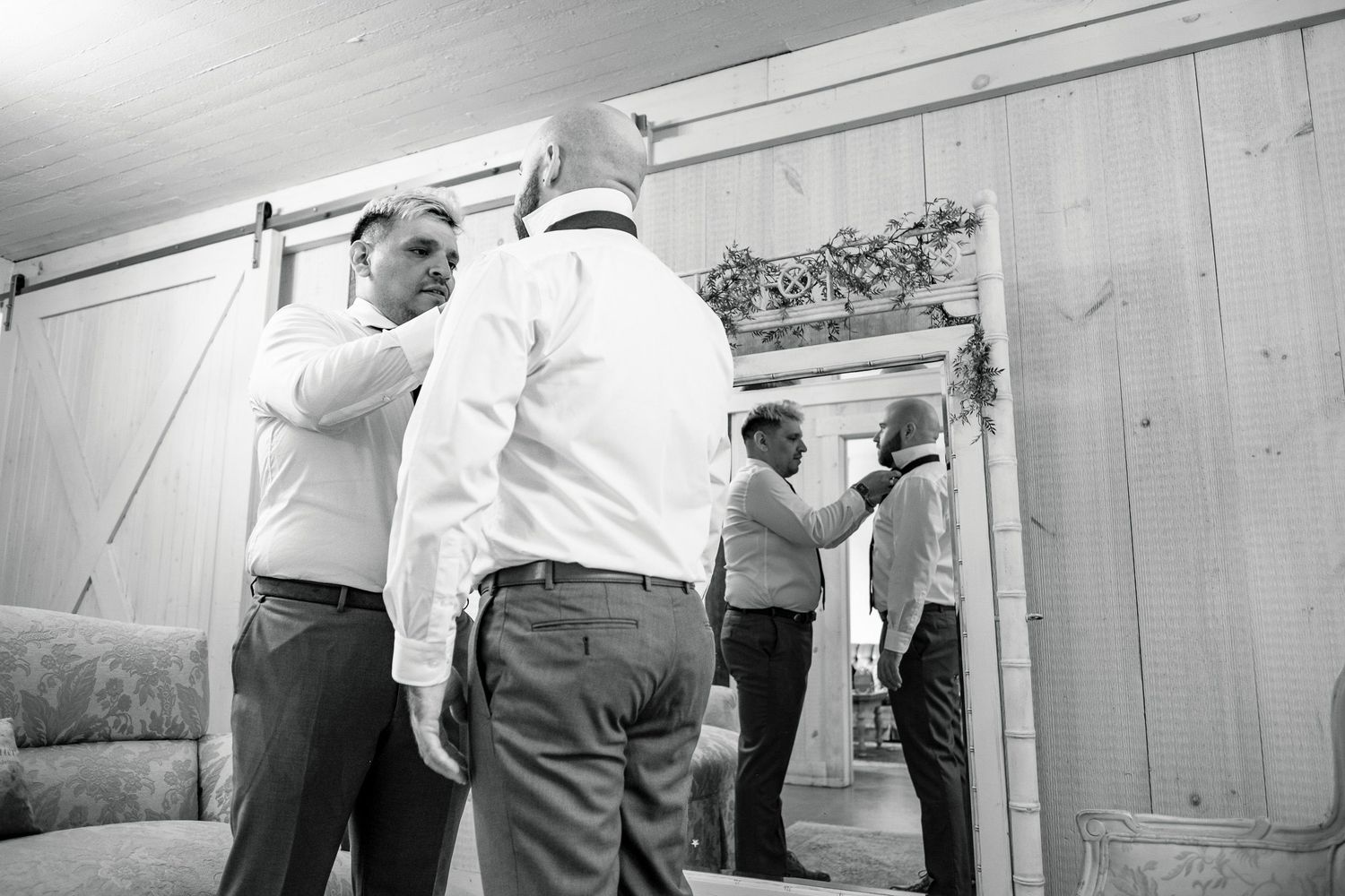 Groomsmen getting ready together near mirror in rustic barn venue, black and white.