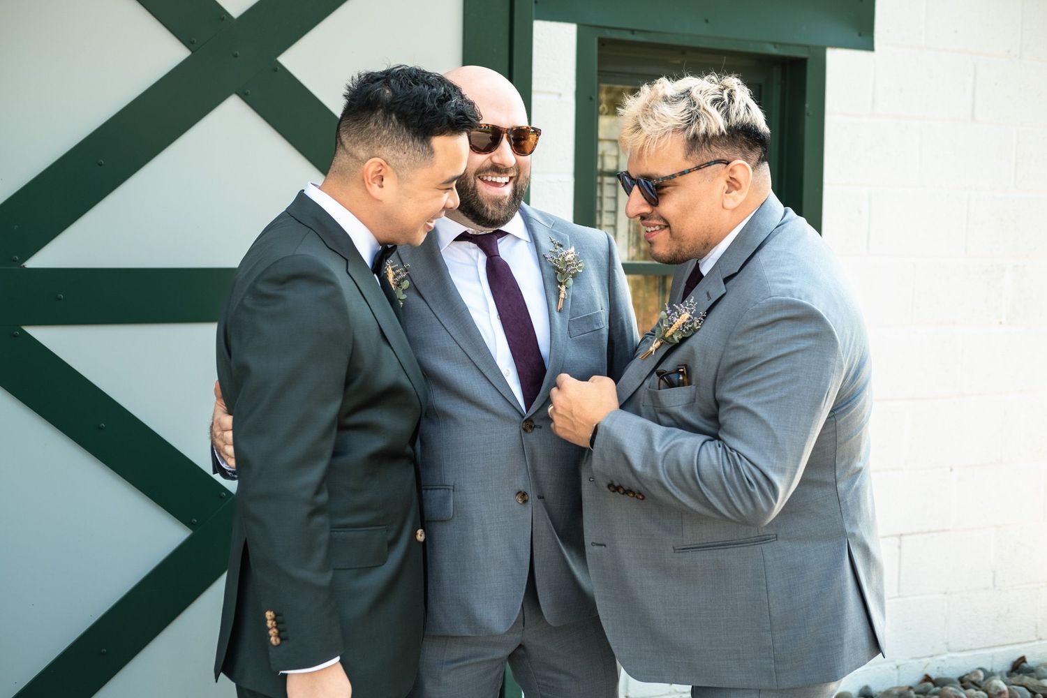 Three men in suits share a joyful moment together outside a white barn wedding venue.