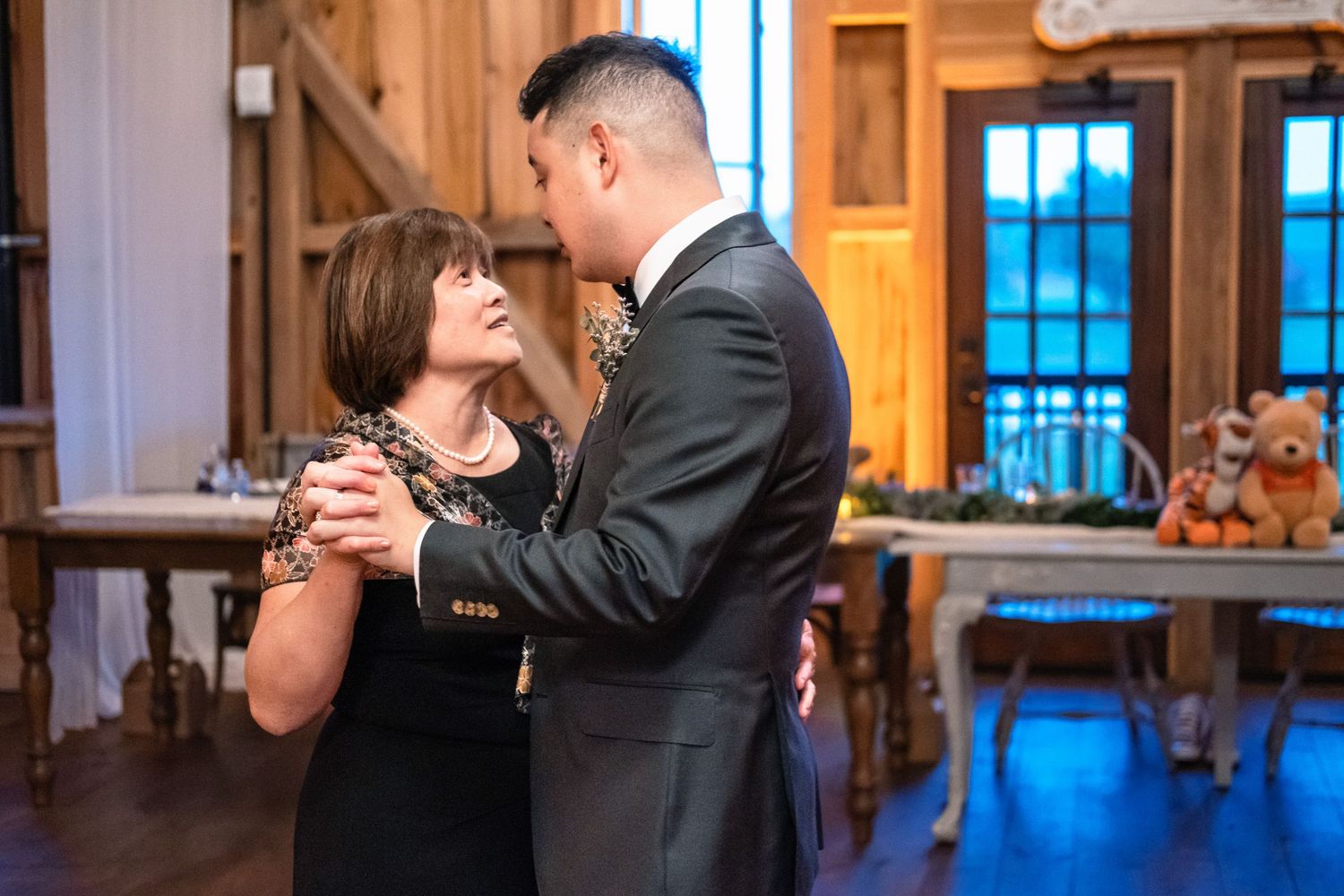 Groom shares a tender mother-son dance inside a warmly lit rustic barn wedding venue.