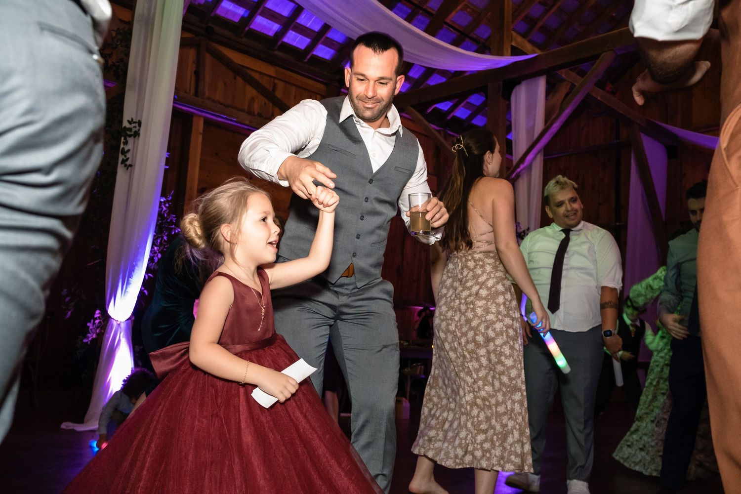 A man in a vest dances with a young flower girl in a burgundy dress at a wedding reception.
