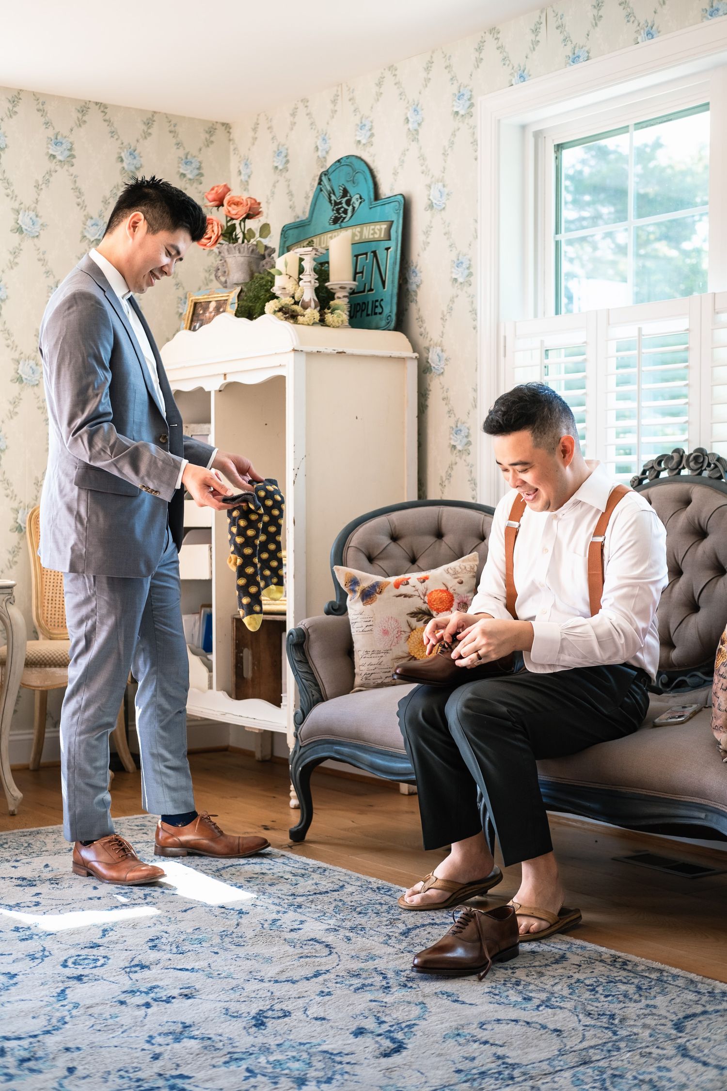 Two groomsmen getting ready in an elegant floral room, one seated putting on shoes.