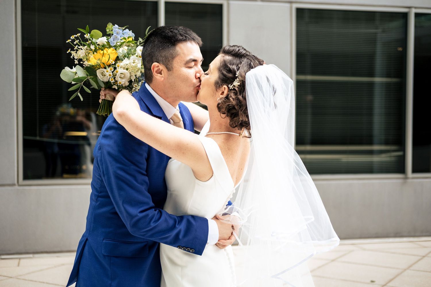 Bride and groom share a romantic kiss outdoors, bride holding yellow and blue floral bouquet.