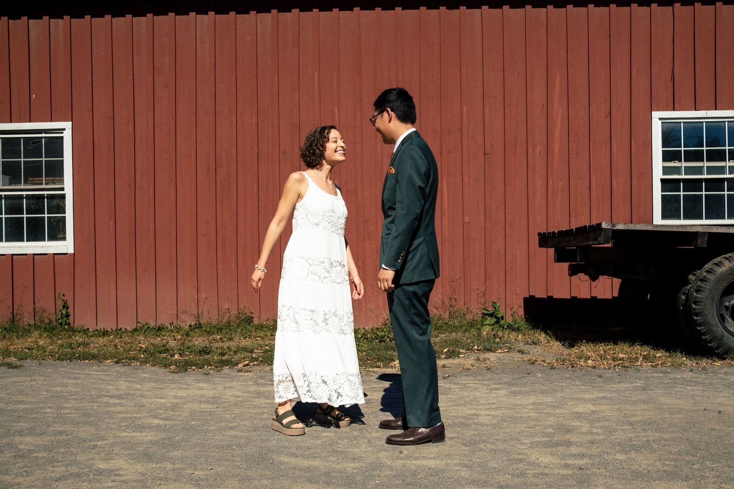 A smiling bride and groom hold hands and face each other outside a classic red barn.