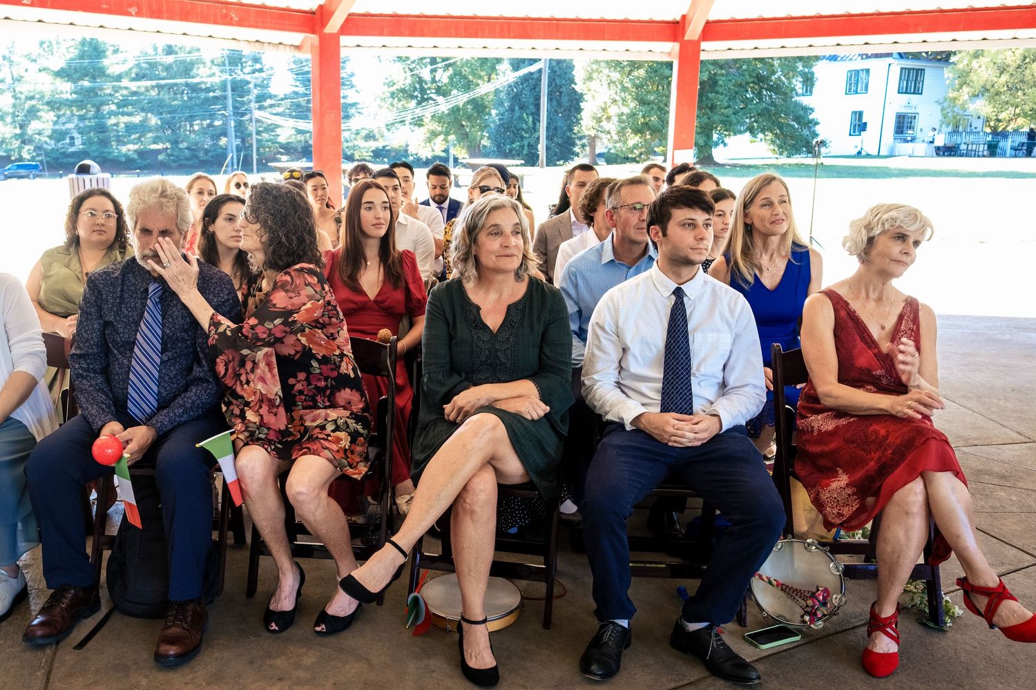 Wedding guests seated outdoors under a pavilion watching a ceremony on a sunny day.