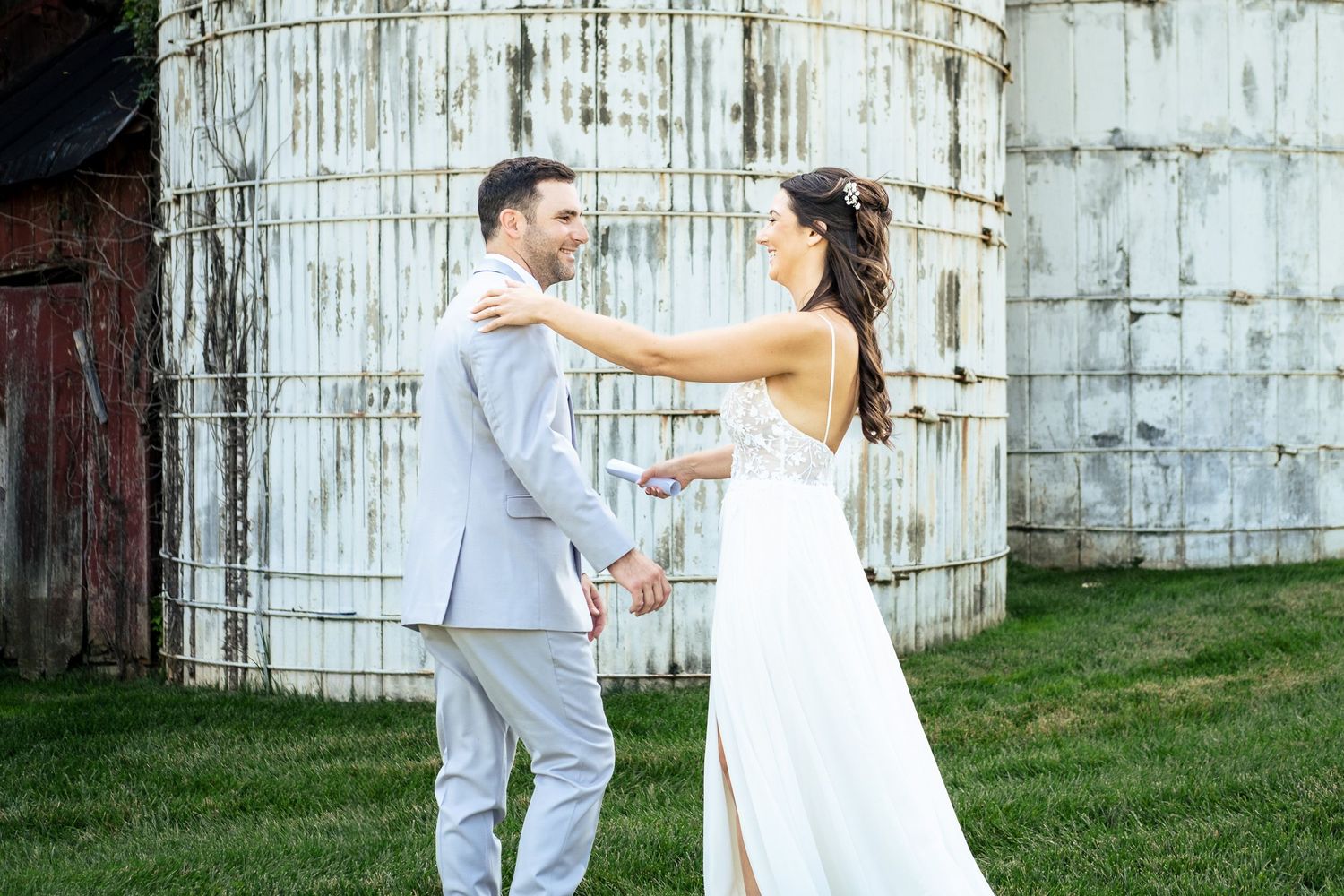 Bride and groom share a joyful first look moment beside vintage weathered grain silos on a farm.