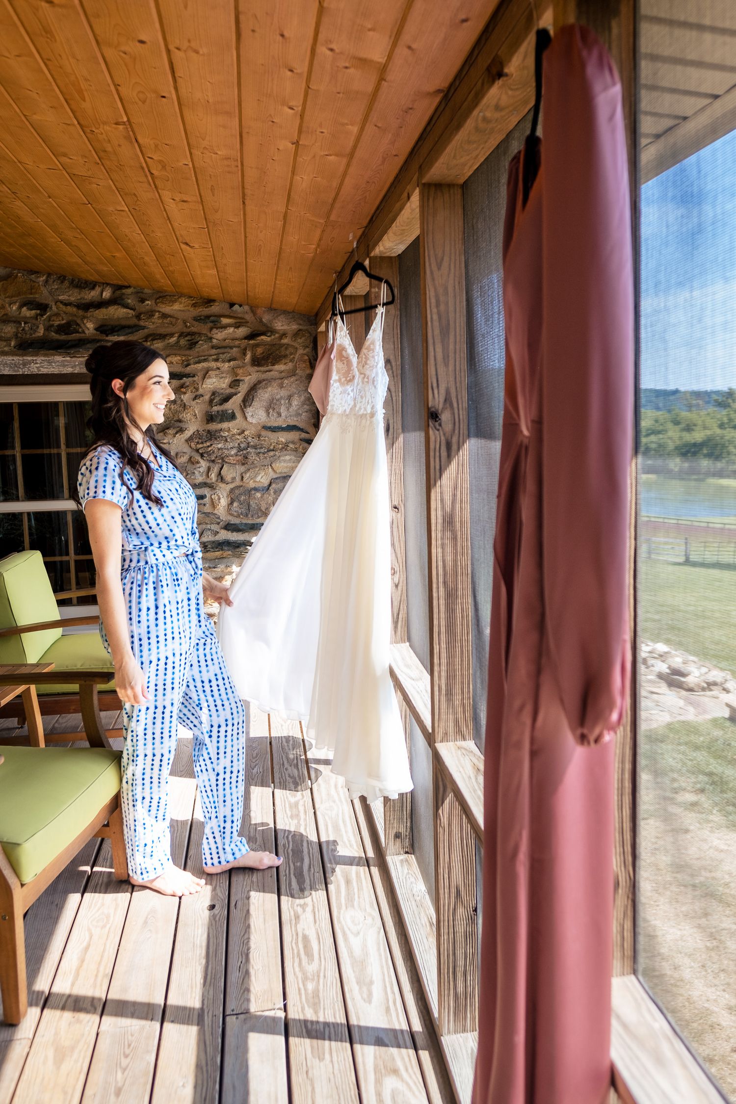 Bride in blue pajamas admires her wedding dress hanging on a rustic wooden porch overlooking a river.