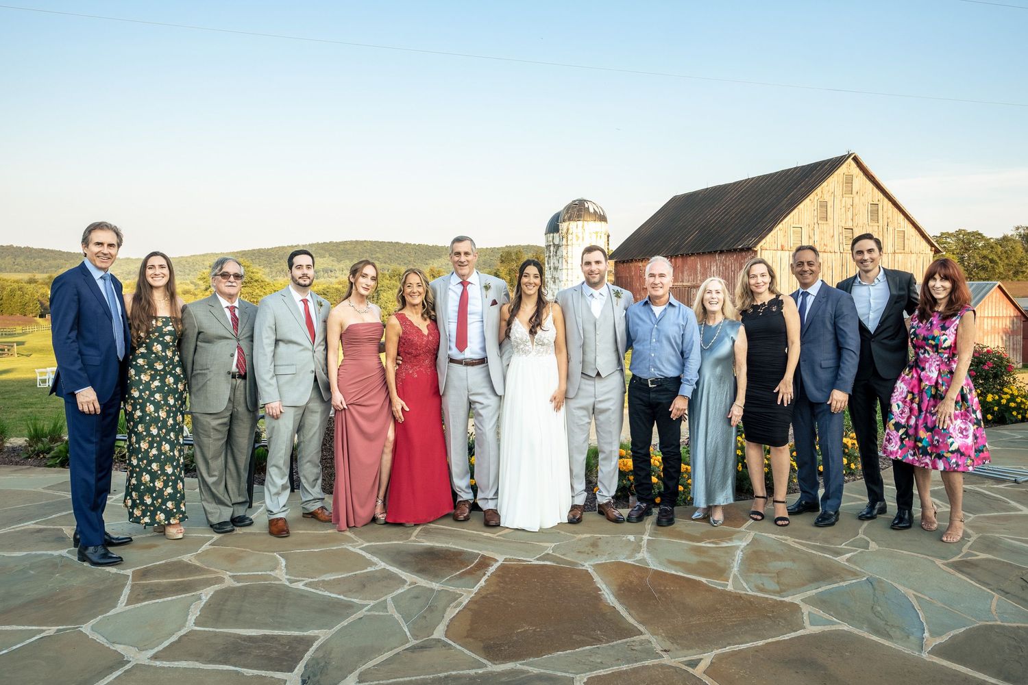 Large wedding family group portrait on stone patio with rustic barn and rolling hills in the background.