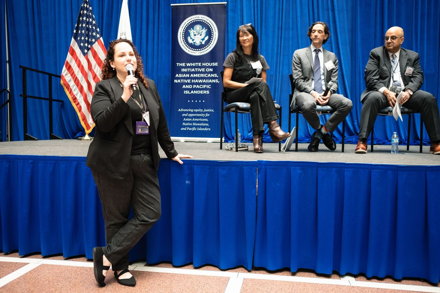 Woman in black suit speaks at White House Initiative panel event with American flag and seated panelists in background.