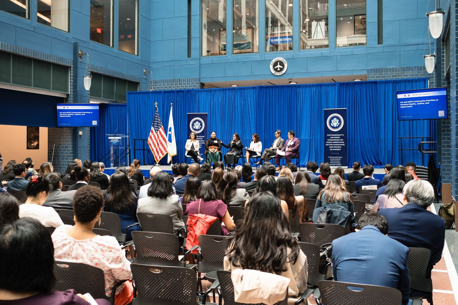 Panel discussion at a government forum with speakers on stage before a large diverse audience, American flag displayed prominently.