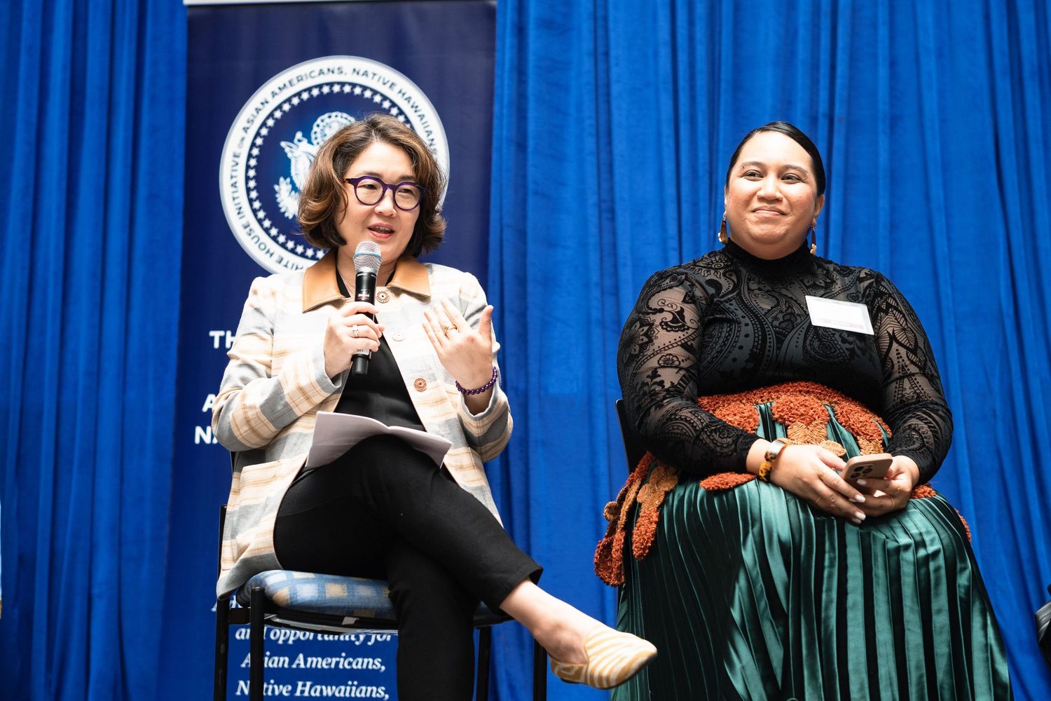 Two women sit on stage during a panel discussion in front of a blue presidential backdrop.