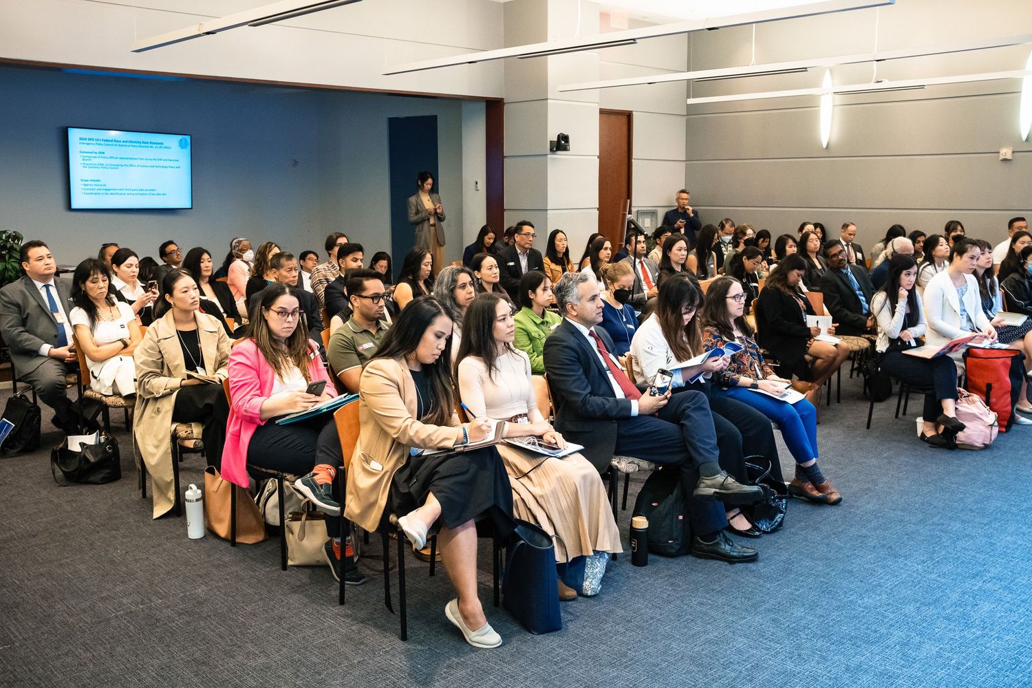 Large audience of diverse professionals seated in rows at a formal conference or seminar in a modern meeting room.