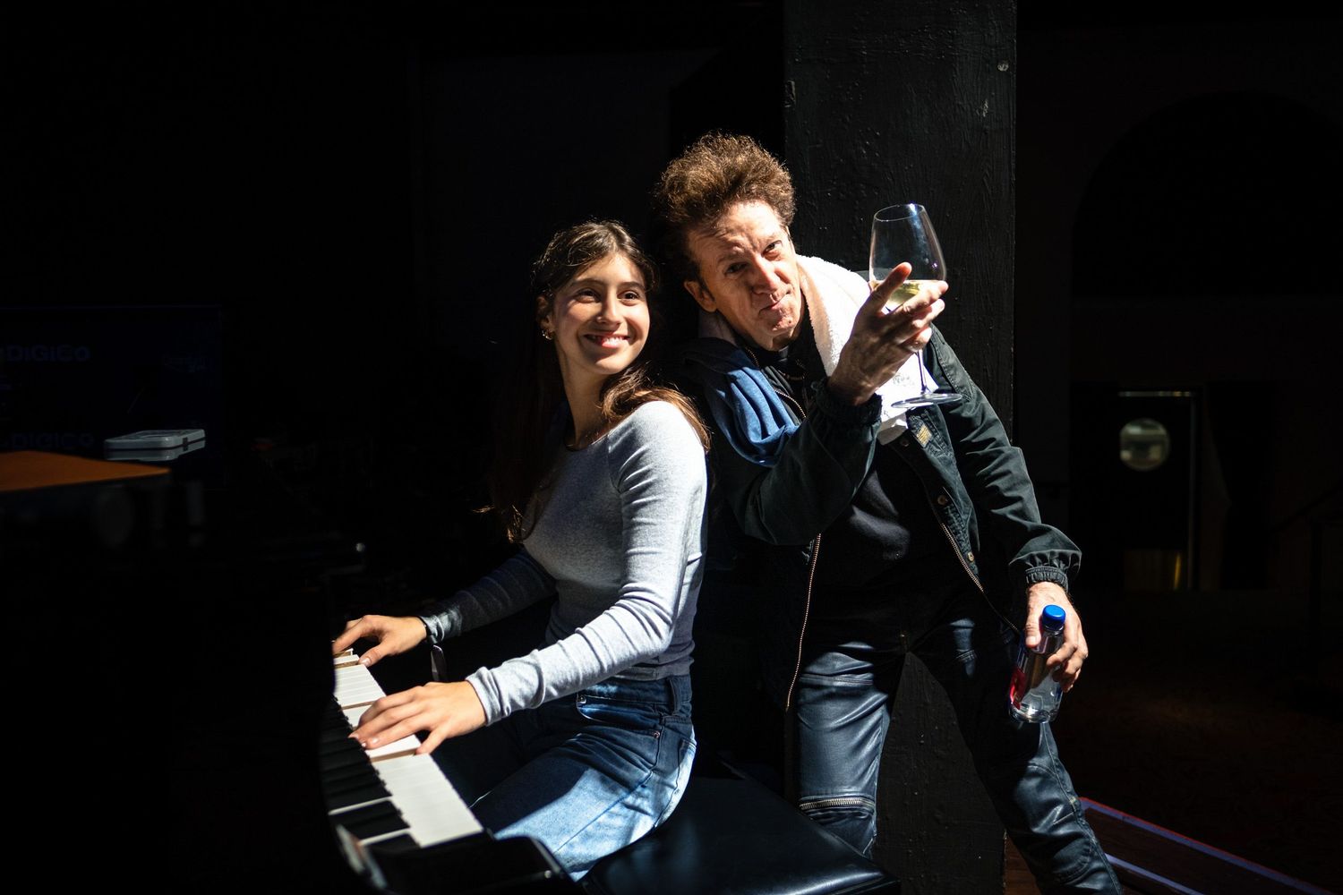 Two musicians pose back-to-back, one playing piano and one holding a microphone, in a dramatic stage setting.