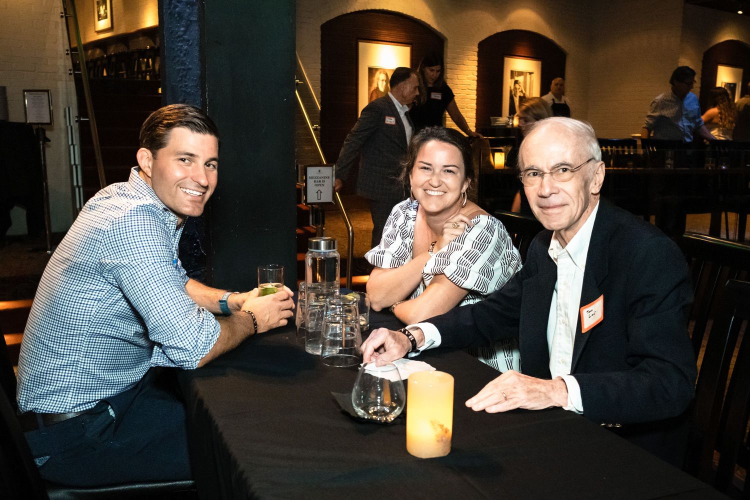 Three people smile and pose at a dark table with drinks at an indoor social gathering event.