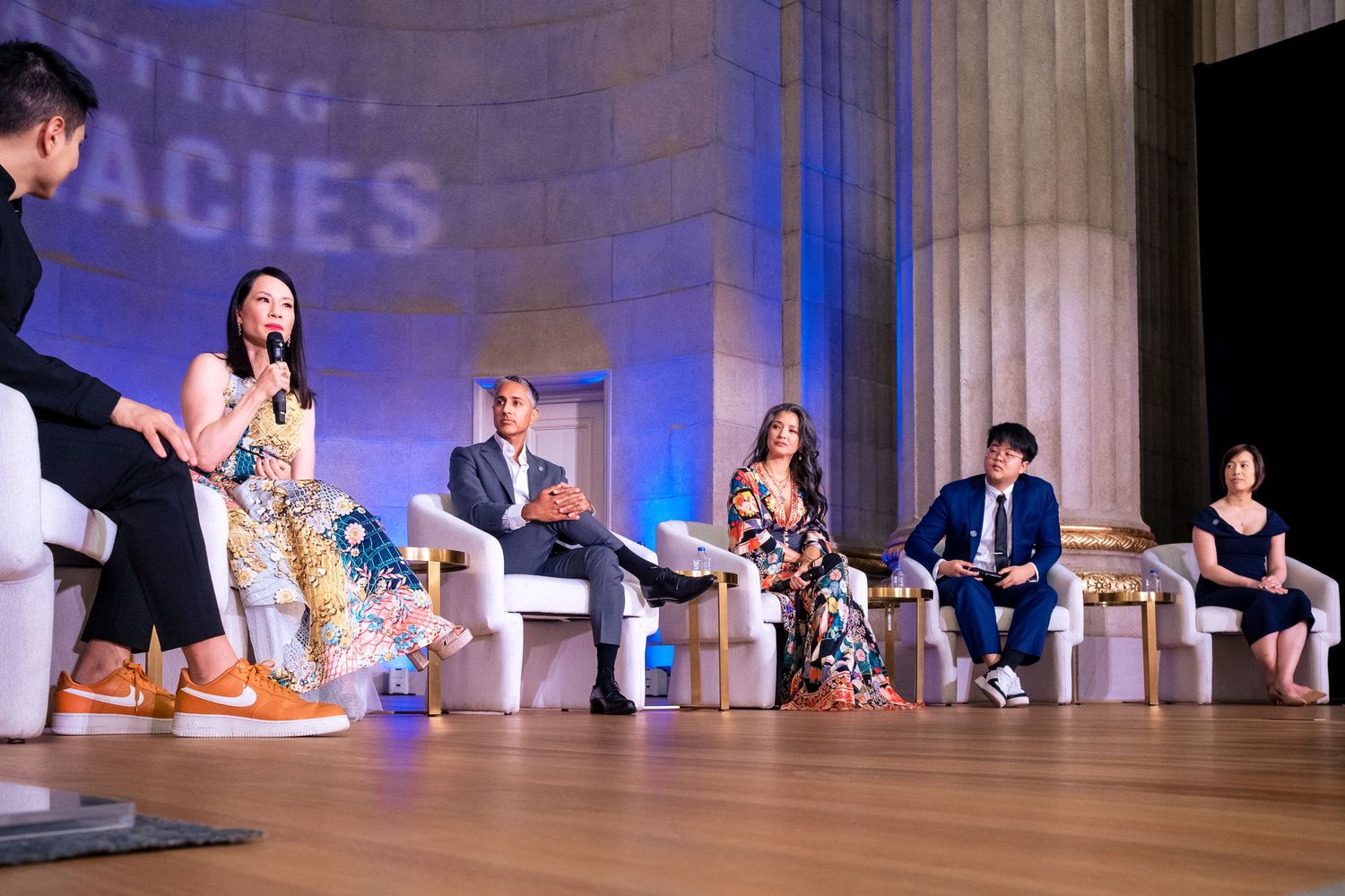 Panel discussion on stage at a formal event with six speakers seated in white chairs before classical columns.