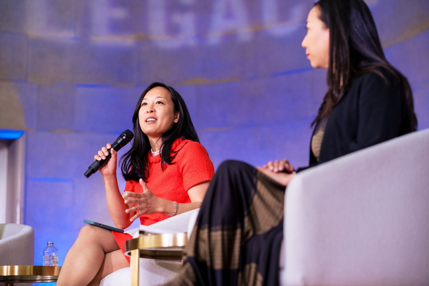 Two women in conversation on stage at a Legacy conference, one in red holding a microphone.