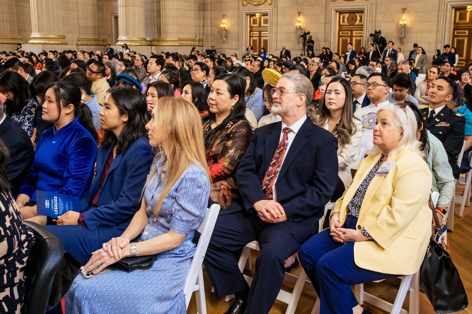 Diverse group of attendees seated at a formal indoor ceremony in a grand hall with white chairs.